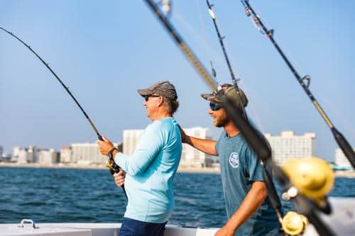 Two people on a boat are fishing with rods in a coastal area. One person assists the other as they reel in the line, with buildings visible in the background.