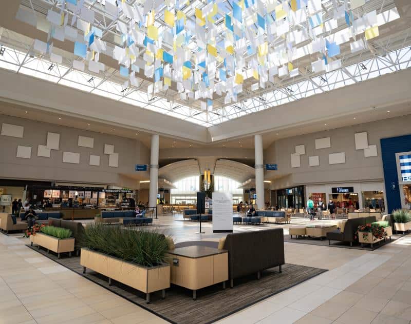 A spacious shopping mall interior with seating areas, plants, and a colorful ceiling installation. Stores and a food court are visible in the background under natural and artificial lighting.