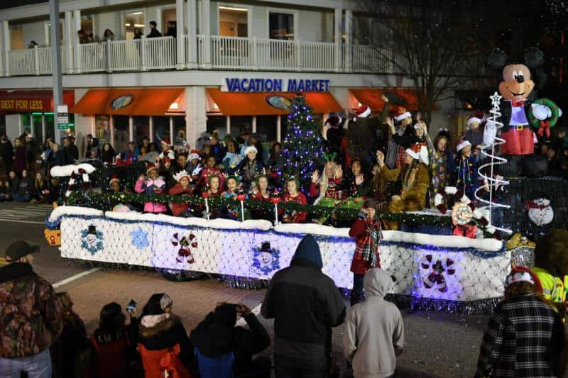 A holiday parade float with people in festive attire and decorations passes by a crowd. There's a large cartoon character figure on the float.