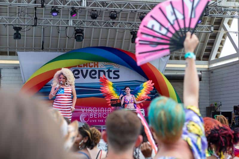 People gathered at an outdoor Pride event. A person on stage with a microphone wears a dress with stars and stripes; another person stands with rainbow wings. Audience members hold various items.