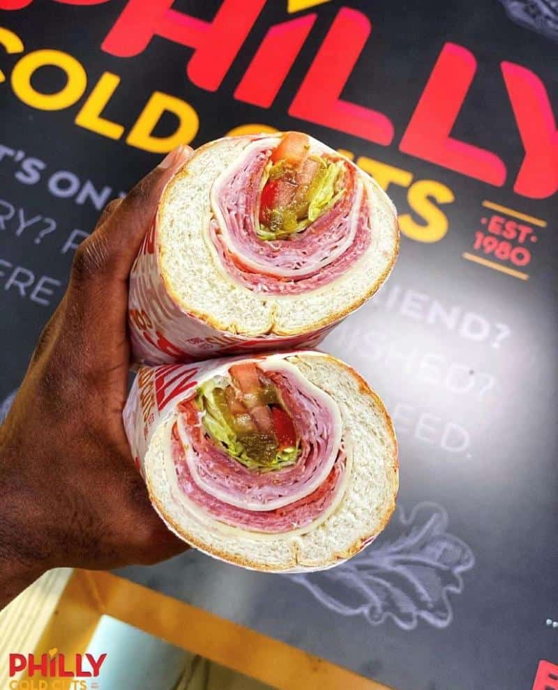 A hand holds a sliced hoagie sandwich filled with layers of meat, cheese, lettuce, pickles, and tomato, against a blurred background with red and yellow text.