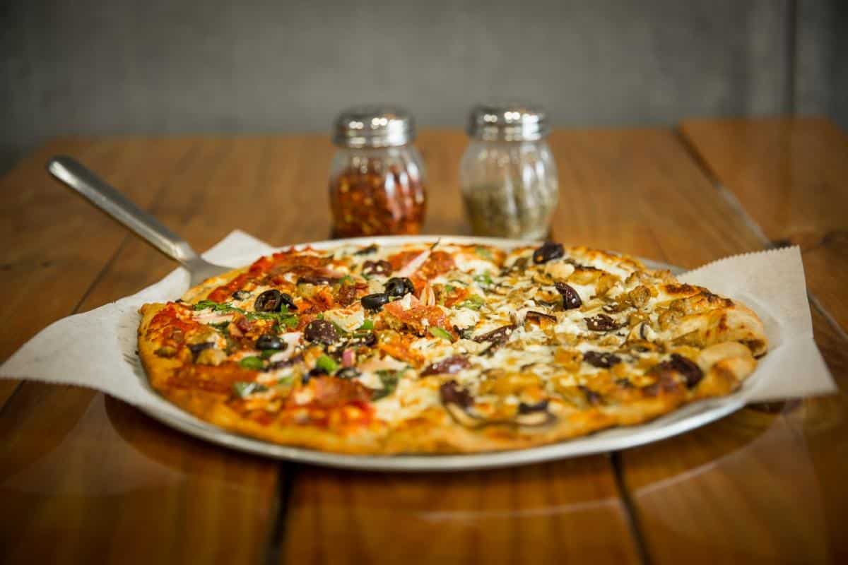 A half-and-half pizza with various toppings on a metal tray, placed on a wooden table. Two spice shakers are in the background.