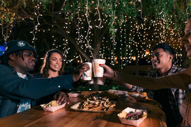 Four people raising cups in a toast at an outdoor table with food, under string lights at night.