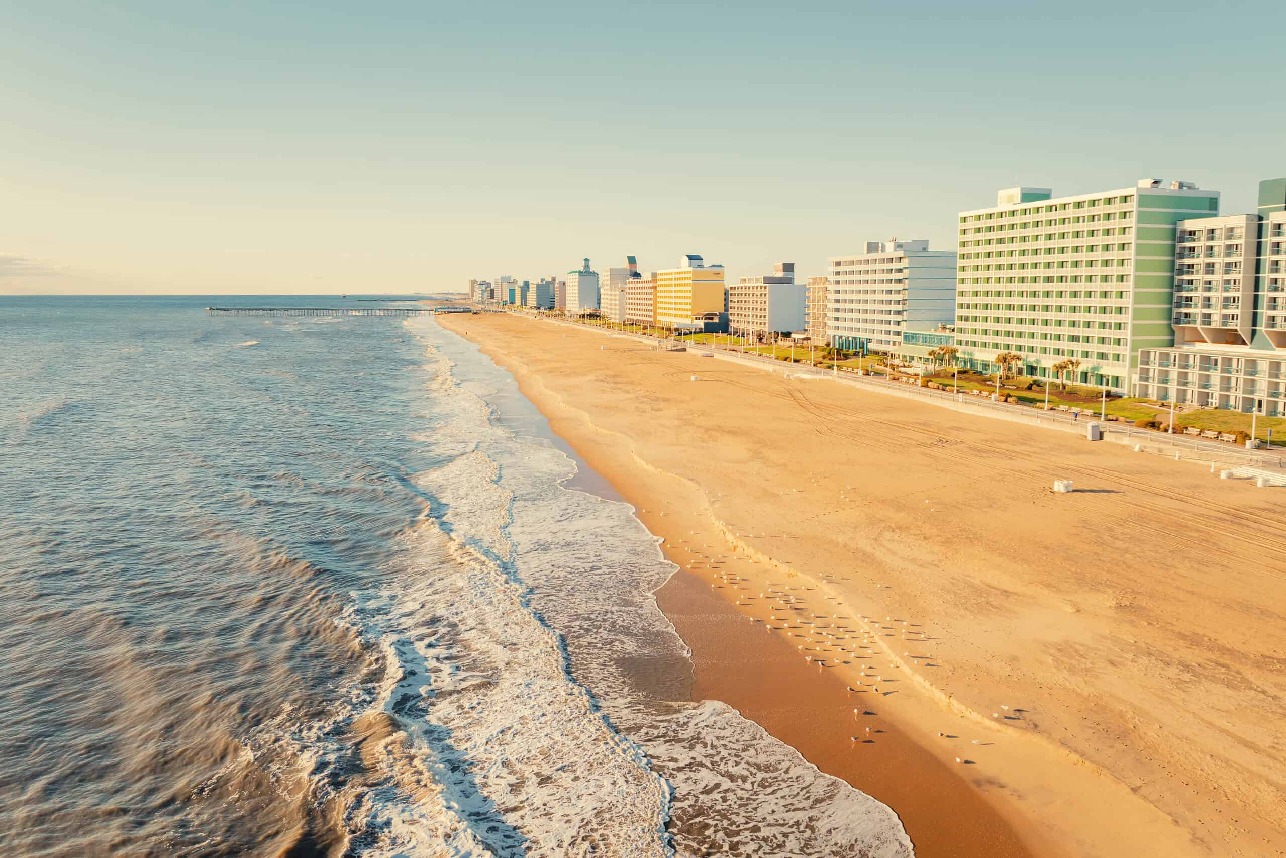 Coastal landscape featuring sandy beach, gentle ocean waves, and modern beachfront buildings under a clear sky.