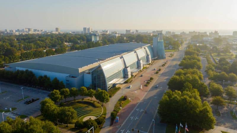 Aerial view of a large, modern convention center with a glass facade, situated beside a tree-lined road in an urban area.