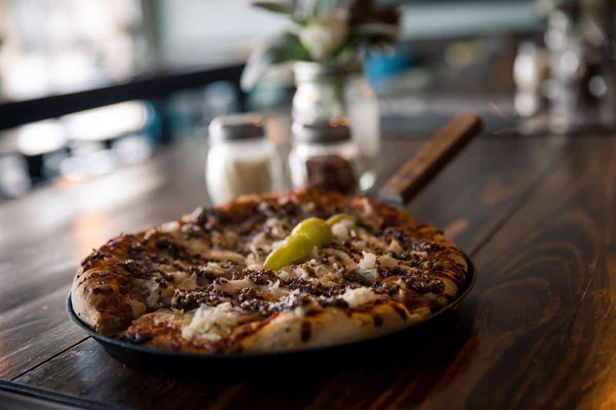 Pizza with minced meat, onions, and a pepper served on a black pan, placed on a wooden table with salt and pepper shakers in the background.