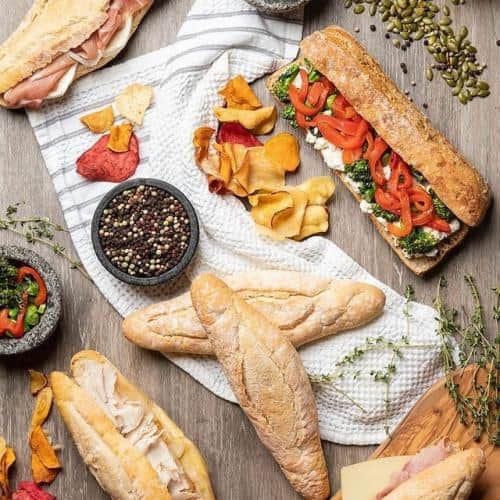 Variety of baguette sandwiches with toppings and chips on a wooden surface, accompanied by a bowl of mixed peppercorns and scattered herbs.