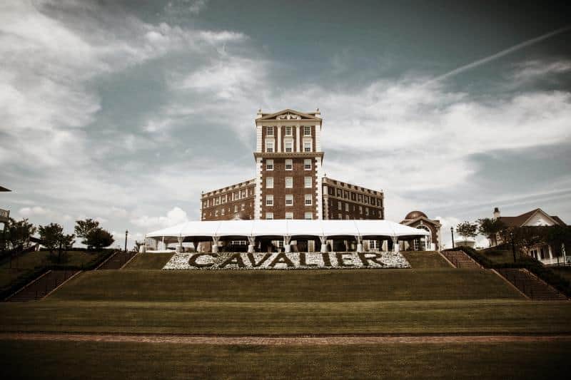 The historic Cavalier Hotel is a multi-story brick building on a grassy hillside with a cloudy sky backdrop.