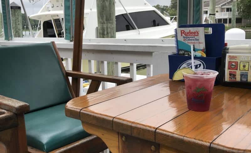 Outdoor table with a magenta drink and a lime wedge beside a green chair. A marina with boats is visible in the background.