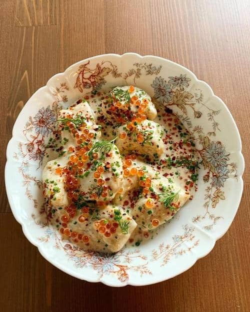 A decorative bowl contains stuffed dumplings topped with red caviar, herbs, and cream sauce, placed on a wooden table.