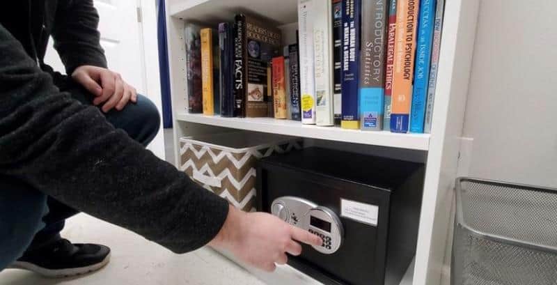 Person kneeling in front of a bookshelf, reaching to open a small safe located on the bottom shelf. Books and a storage box are also visible on the shelf.