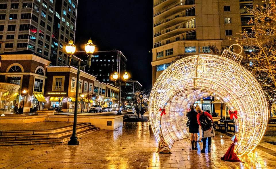 Two people stand under a lit decorative arch in a city plaza at night, surrounded by tall buildings and illuminated streetlights.