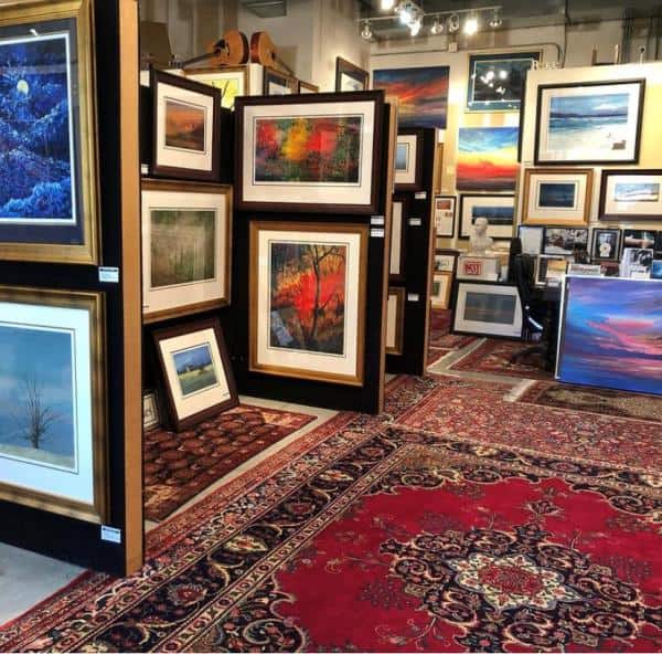 Art gallery interior with framed paintings displayed on walls and stands. The floor is covered with red Persian-style rugs.