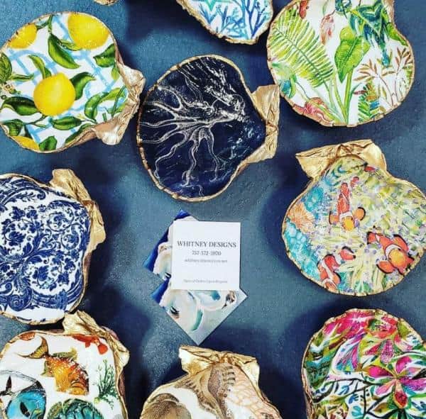 Decorative oyster shells with colorful designs, including citrus fruits, ocean scenes, and floral patterns, displayed around a business card on a blue surface.