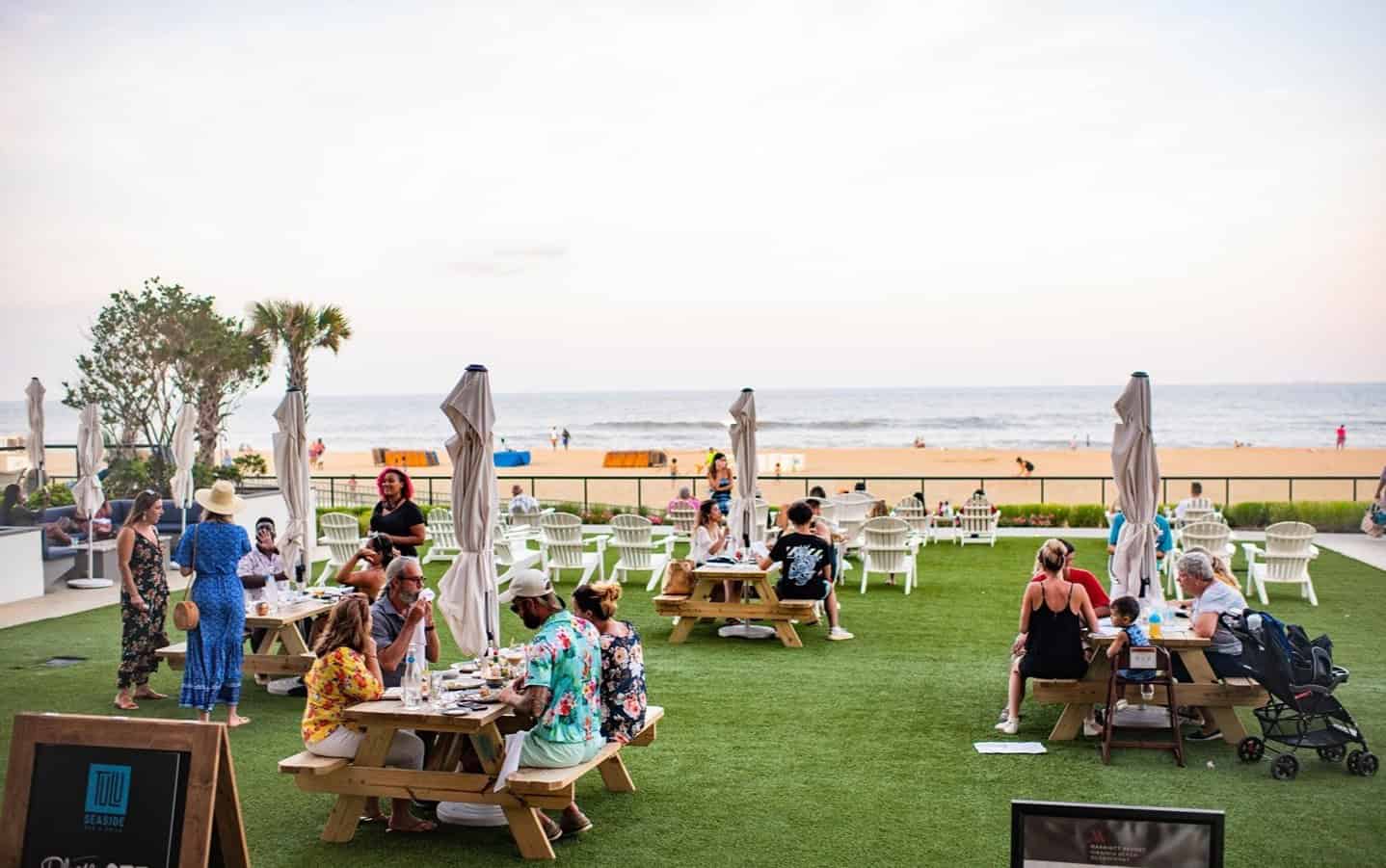 People sitting and socializing at picnic tables on a grassy area near the beach. The ocean is visible in the background under a cloudy sky.