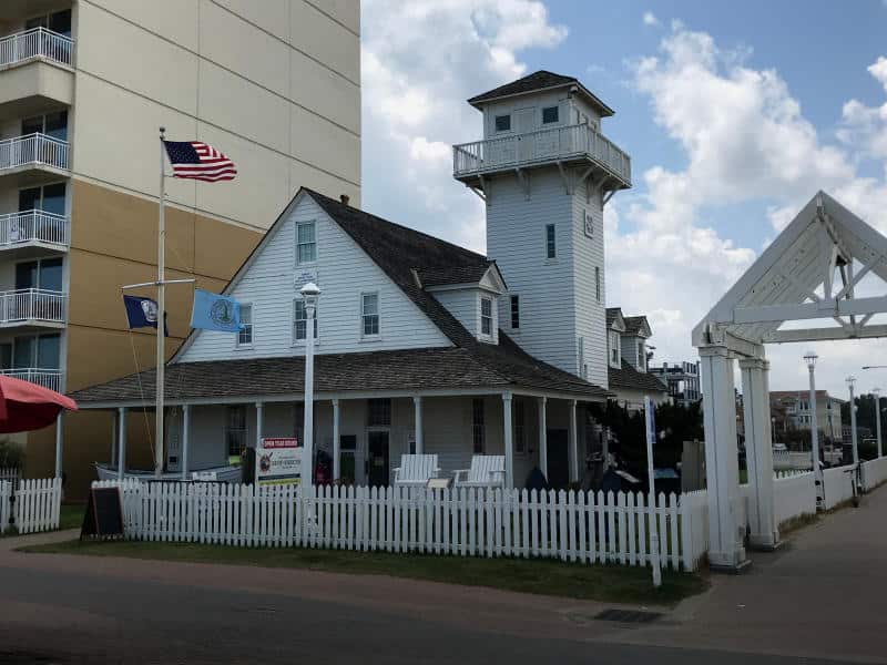 The Surf & Rescue Museum is a white building with a tower and picket fence, surrounded by flags. Adjacent to a taller structure, under a partly cloudy sky.