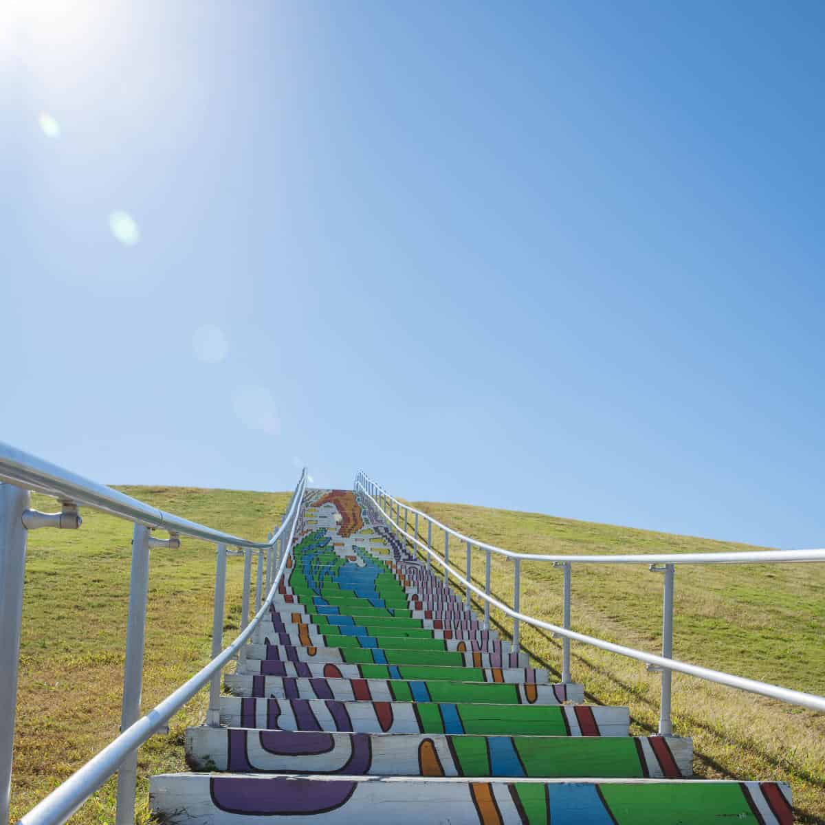 Colorful, painted outdoor stairs with metal railings on both sides ascending a grassy hill under a clear blue sky.