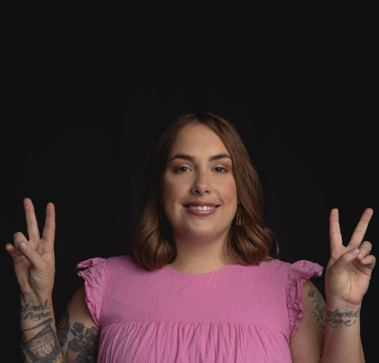 A person in a pink blouse holds up both hands showing peace signs, with tattoos visible on their arms, against a dark background.