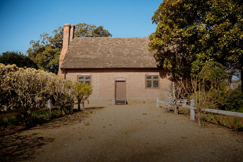 The Adam Thoroughgood House is a small brick house with a steep roof, central door, and two windows. Surrounding foliage and a clear blue sky are visible.