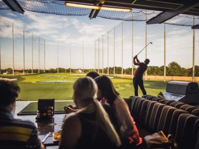 People sitting at a table inside a golf entertainment venue as a person swings a golf club. Targets are visible on the outdoor range.