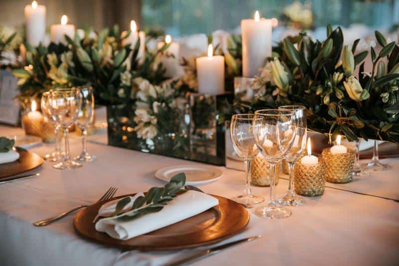 Elegant table setting with white linens, gold chargers, wine glasses, and folded napkins. Floral centerpieces and lit candles create an ambient atmosphere.
