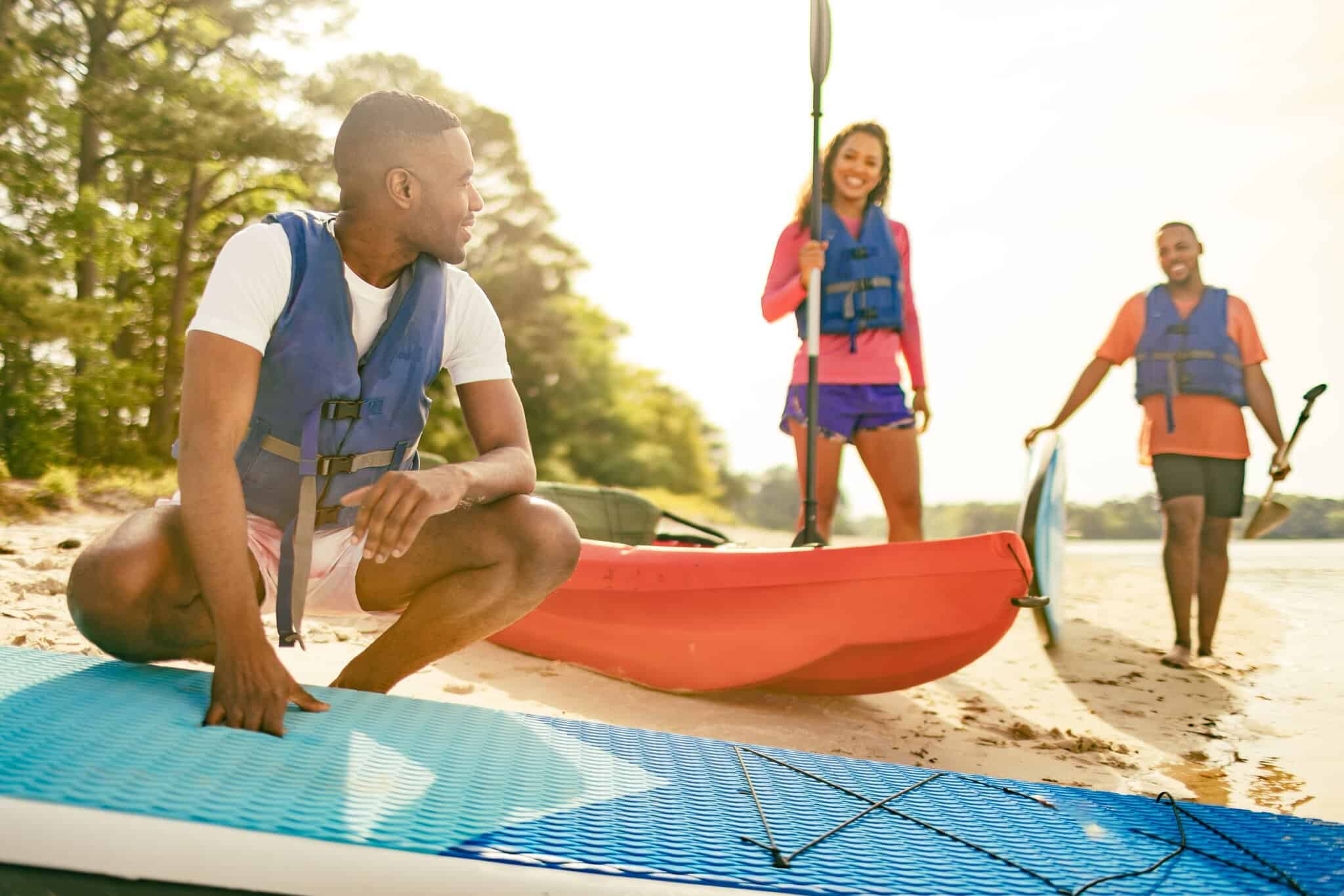 Three people wearing life jackets prepare for water activities on a sandy beach at The Narrows, with a kayak and paddleboards beside them on a sunny day.