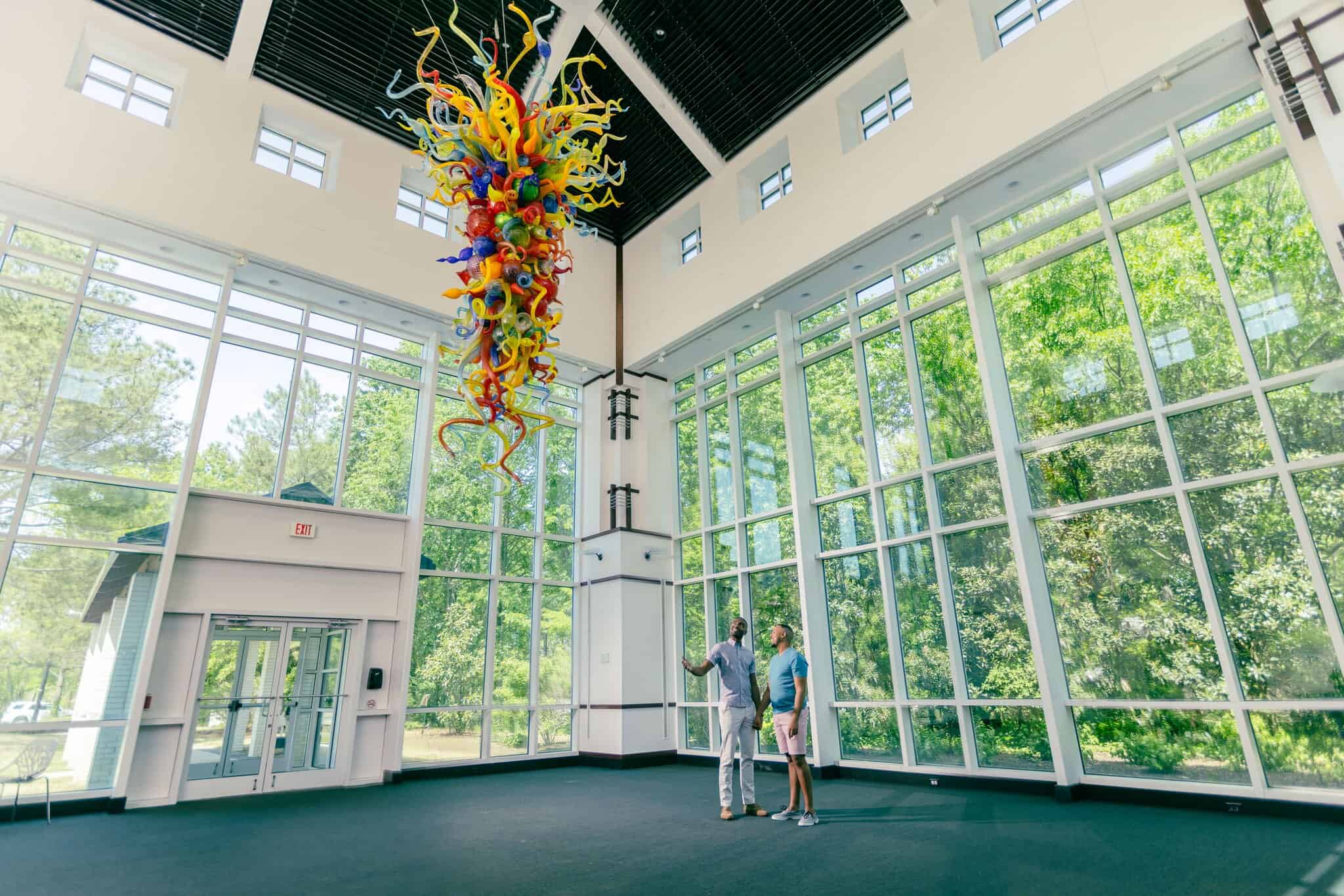 Two people stand in a spacious, sunlit room with large windows, looking up at a colorful glass sculpture by Dale Chihuly hanging from the ceiling at the Virginia Museum of Contemporary Art.