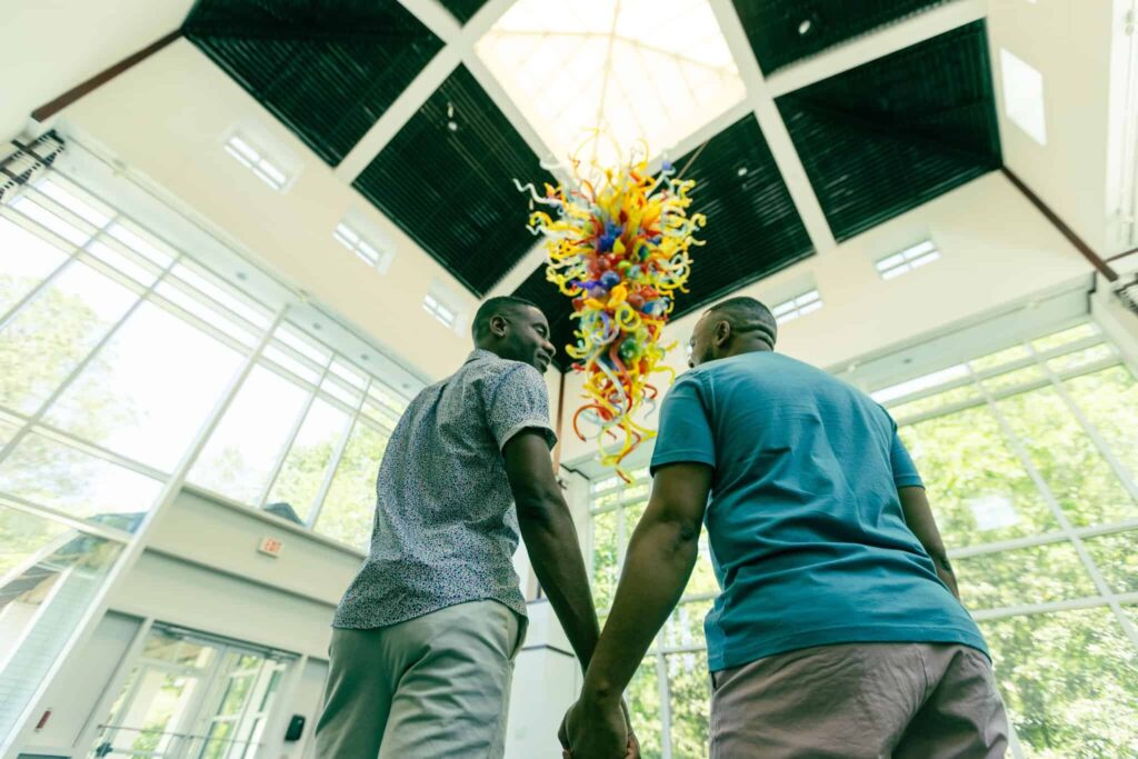 Two people holding hands stand indoors beneath a large, colorful glass sculpture by Dale Chihuly suspended from a high, sunlit ceiling at the Virginia Museum of Contemporary Art.