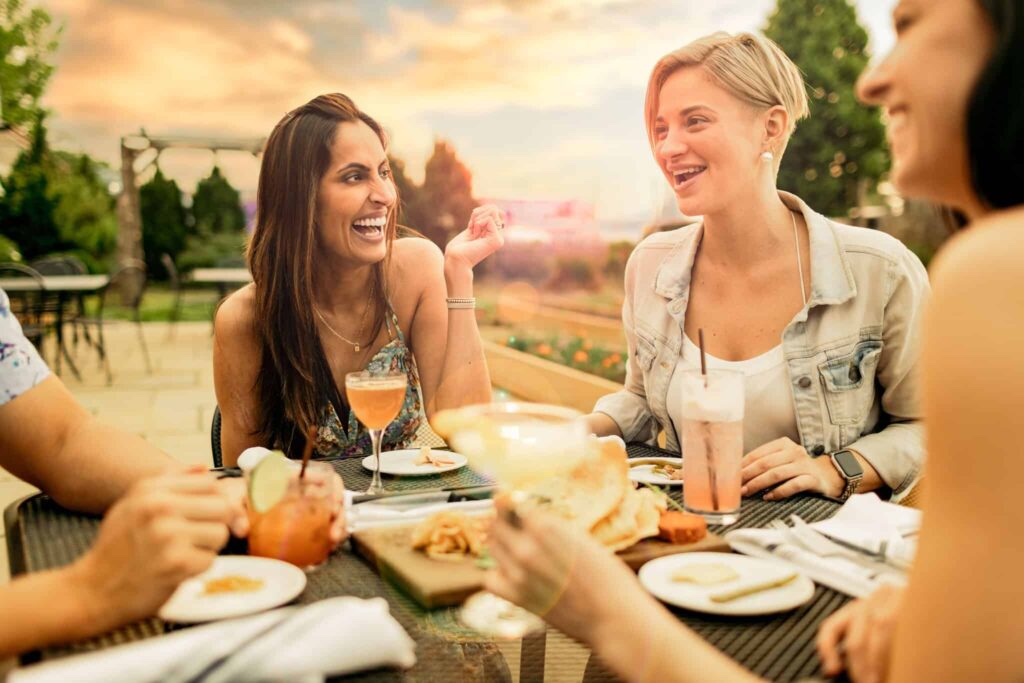 Four people sit at an outdoor table, laughing and enjoying drinks and food in a garden setting during sunset.