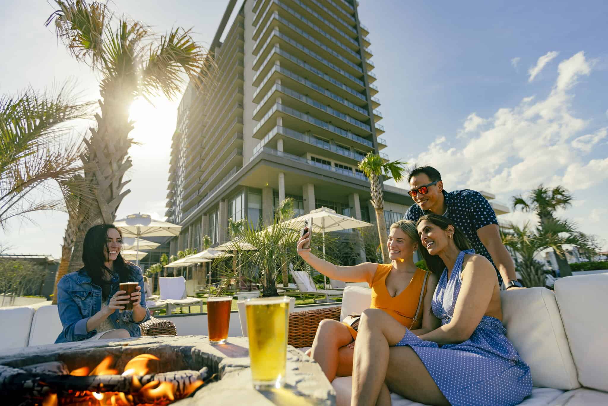 Four people sit around an outdoor fire pit with drinks, taking selfies in front of a modern high-rise building on a sunny day.
