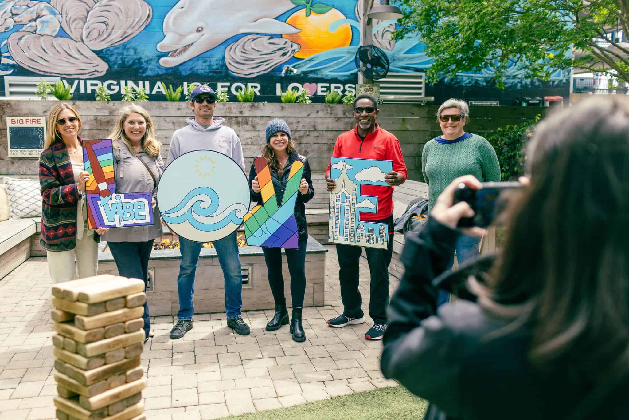Six people stand outdoors holding colorful letters spelling "VIBE" as another person takes their photo; a mural and trees are in the background.