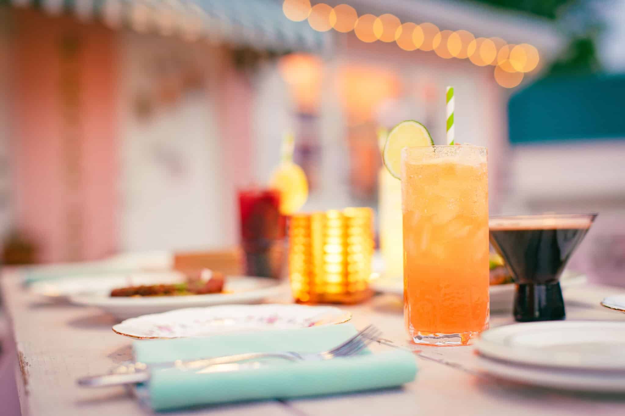 A close-up of a table set with plates, cutlery, and drinks at the Pink Dinghy restaurant including an orange cocktail with ice and a striped straw, outdoors with blurred lights in the background.