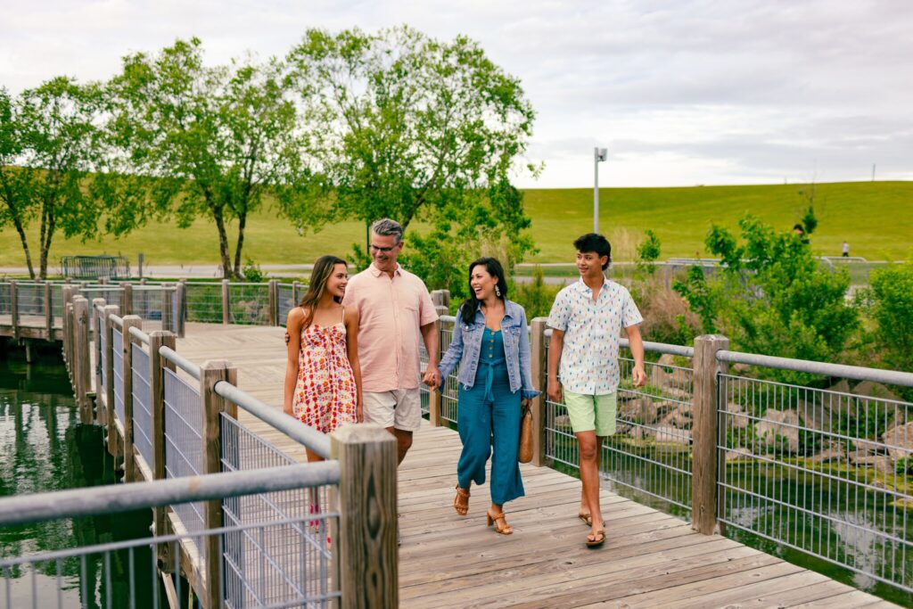 Four people walk together on a wooden boardwalk over water, surrounded by trees and grassy hills, on a partly cloudy day.