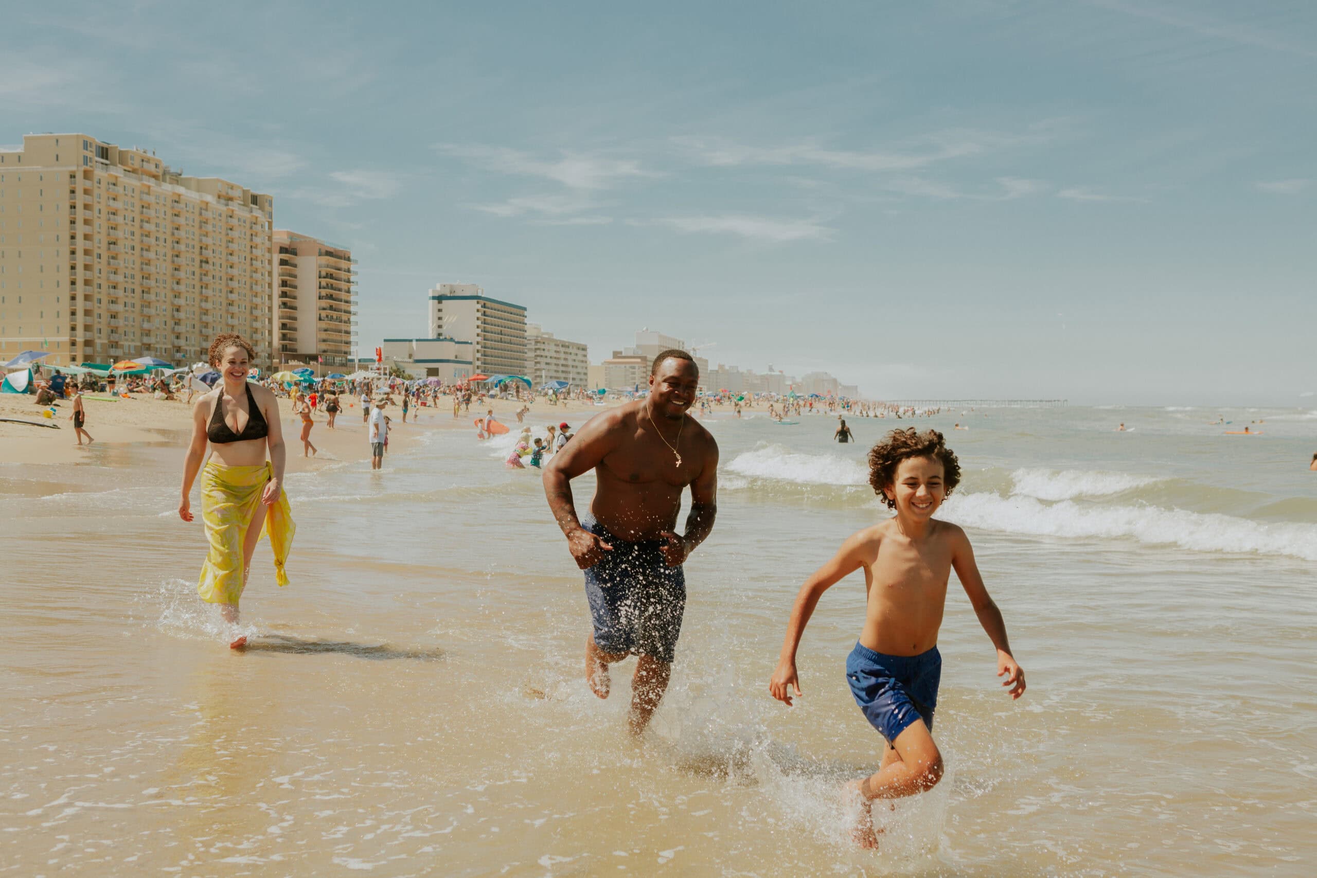 A man and a boy run through the waves at a beach, while a woman walks nearby; hotels, apartments and umbrellas are in the background.