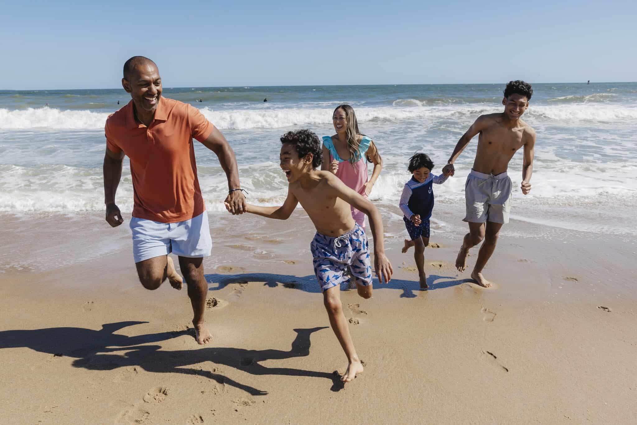 Five people, including three children and two adults, run and smile together on a sandy beach with ocean waves in the background under a clear sky.