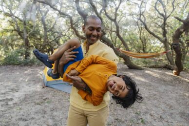 An adult holding a smiling child sideways outdoors at a campsite, with trees, a tent, and a hammock visible in the background.
