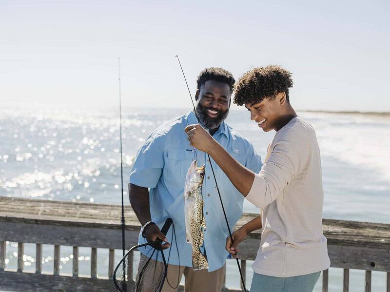 Two people stand on a pier by the ocean, smiling as one holds up a fishing rod with a fish they caught; a fishing net is visible.