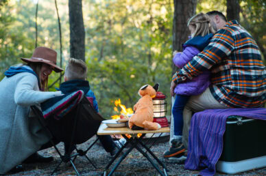 A family enjoys a camping trip around a fire, with a mother and son in chairs, while a father hugs his daughter, featuring a plush fox on a table.