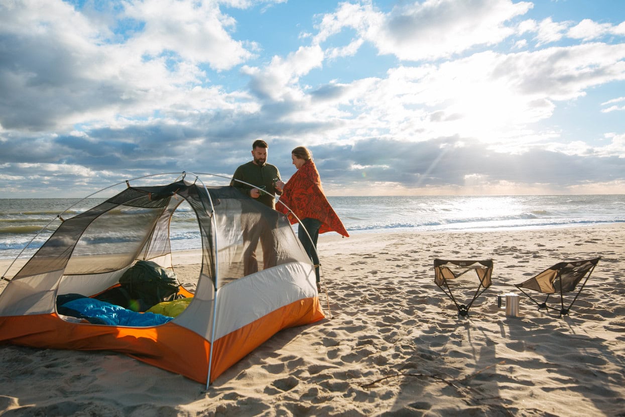 A man and woman set up a tent on the beach, with chairs and a scenic ocean backdrop under cloudy skies.