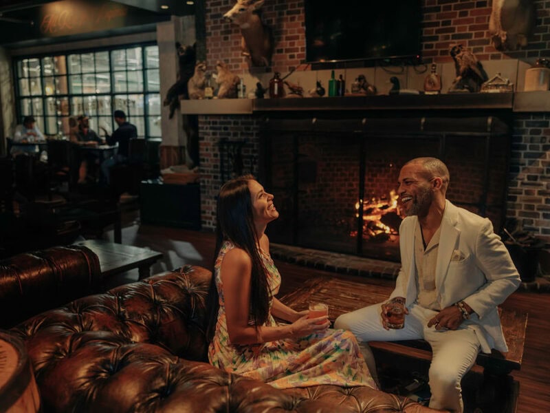 A stylish couple enjoys drinks by a cozy fireplace in a warmly lit lounge, featuring rustic decor and a vibrant atmosphere.