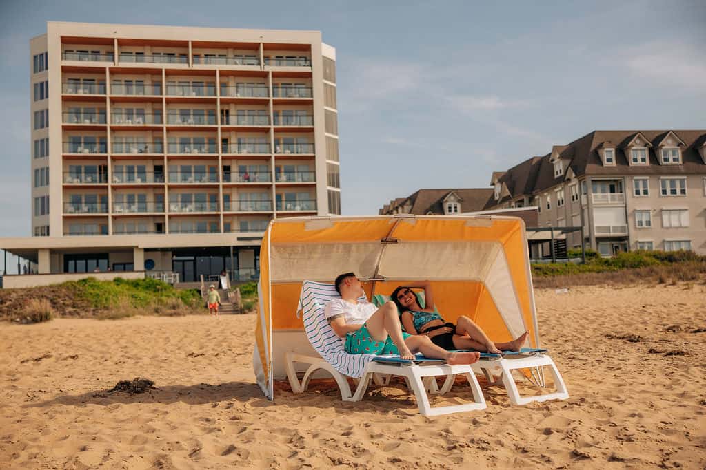 A couple relaxes under a beach cabana on sandy shores, with a modern hotel in the background.
