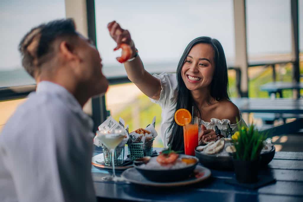 A smiling woman feeds a piece of shrimp to a man during a seaside meal, surrounded by delicious seafood and drinks.