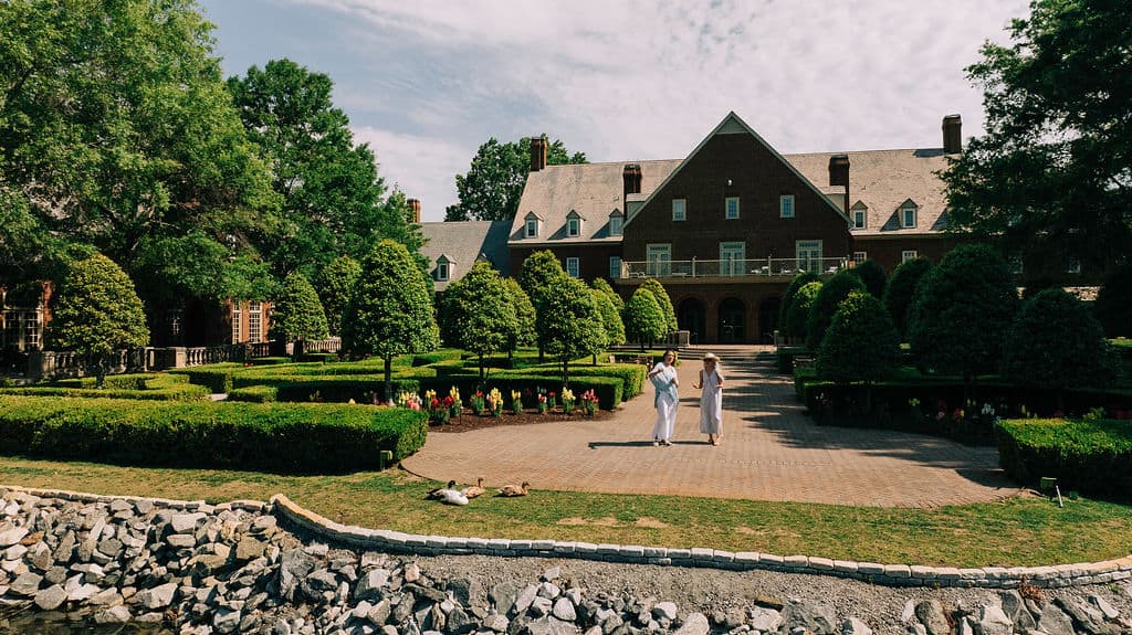 Two women walk across a beautifully landscaped courtyard in front of a large historic mansion, the Founders Inn, surrounded by greenery and flowers.