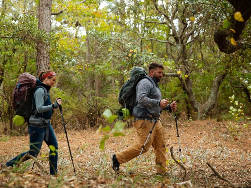 A woman and man hiking through a wooded area, equipped with backpacks and trekking poles.