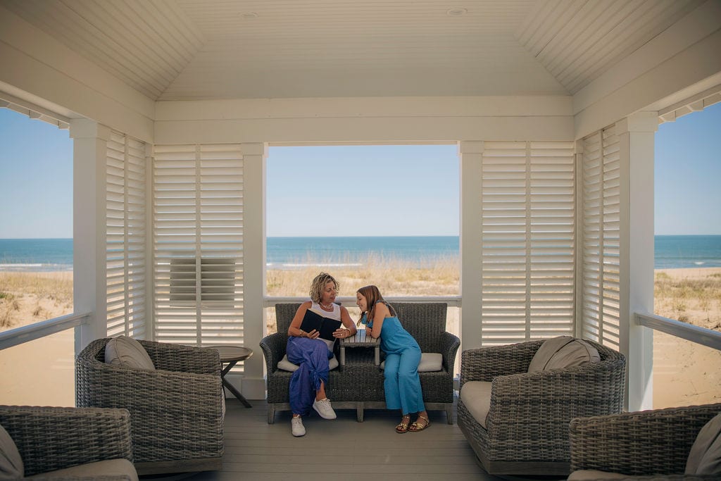 A mother and daughter share a quiet moment on a porch overlooking the beach, seated on comfortable wicker chairs.