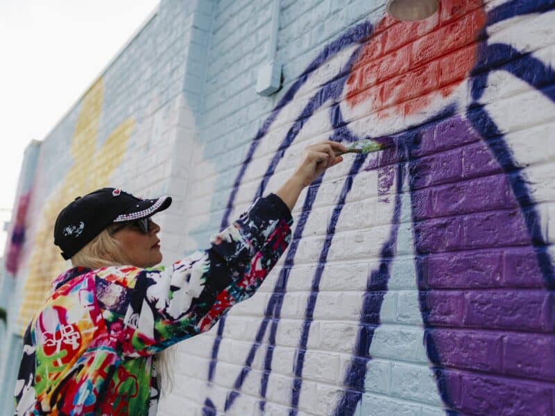 A woman with blonde hair paints a colorful mural on a brick wall, using a brush to add purple paint.