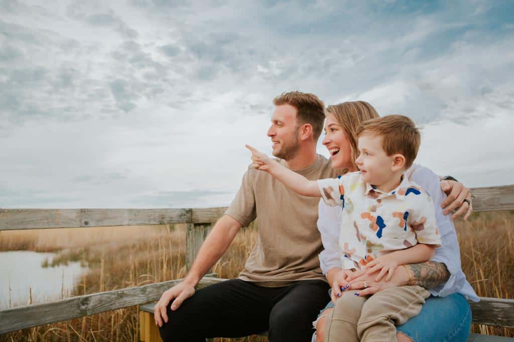 A happy family of three sitting on a wooden railing by a marsh, with the child excitedly pointing at something in the distance.