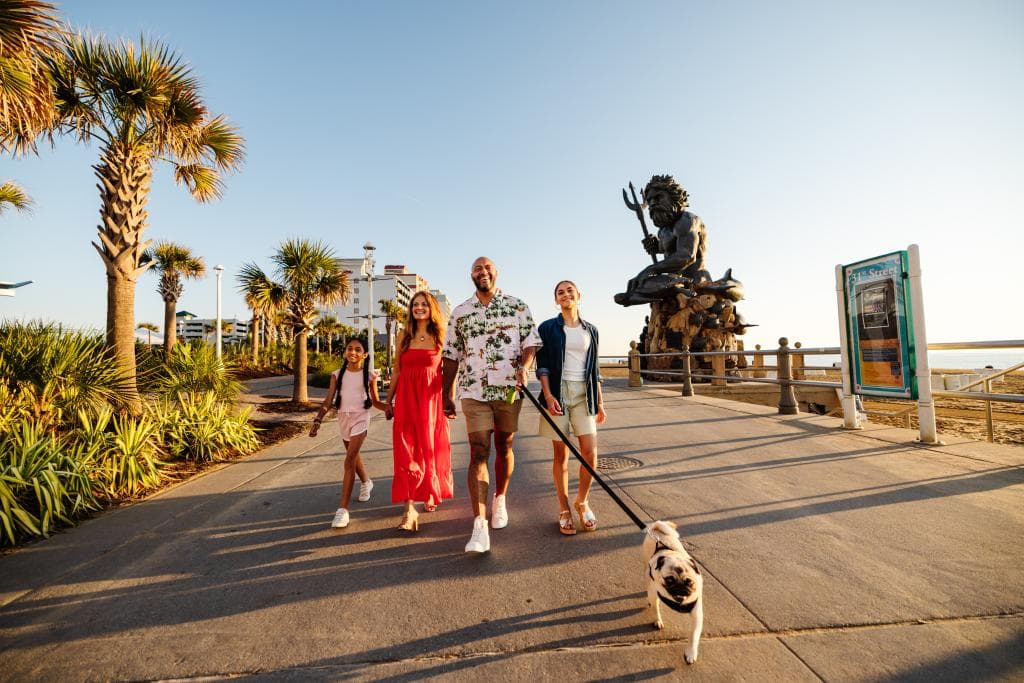 A family with two adults and two children walk along a palm-lined boardwalk, a dog on a leash, with the King Neptune statue in the background.