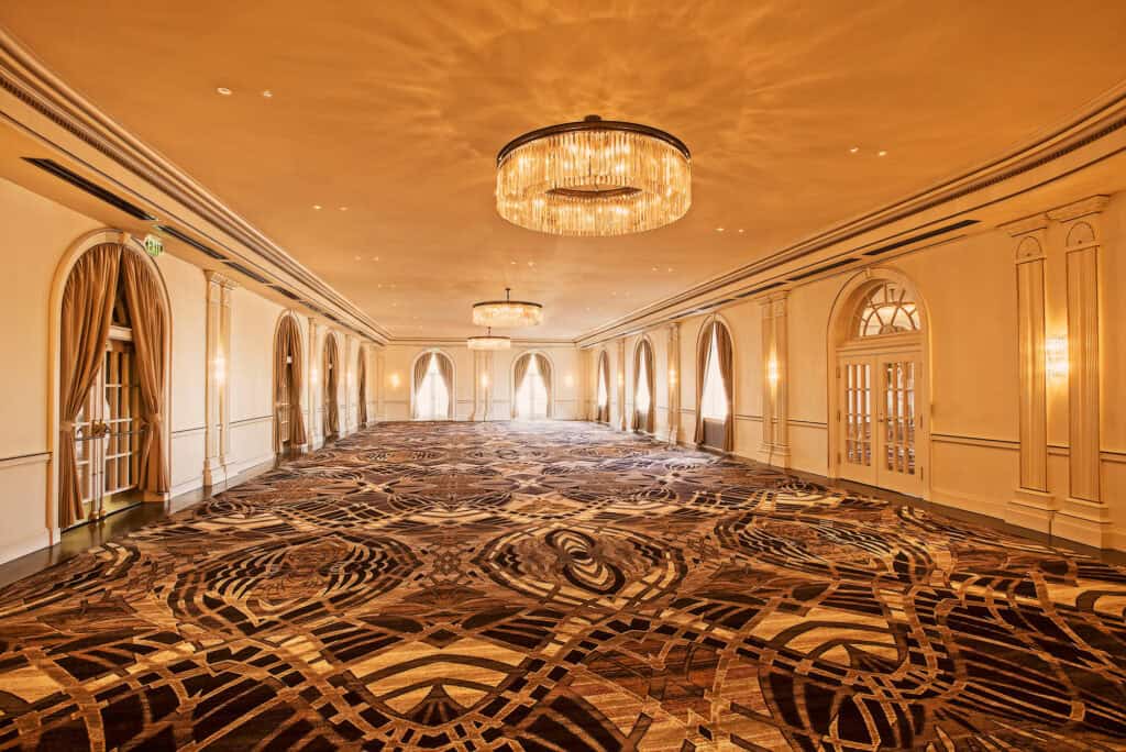 The Crystal Ballroom at The Cavalier Hotel. A spacious event room with elegant chandeliers, ornate carpeting, and large windows, ideal for gatherings and celebrations.