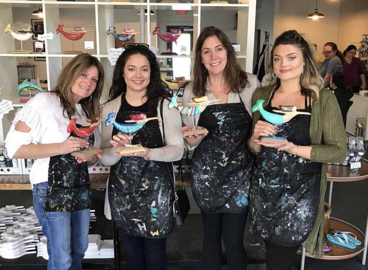 Four women proudly display their painted bird sculptures in a creative studio setting, wearing aprons covered in paint splatters.